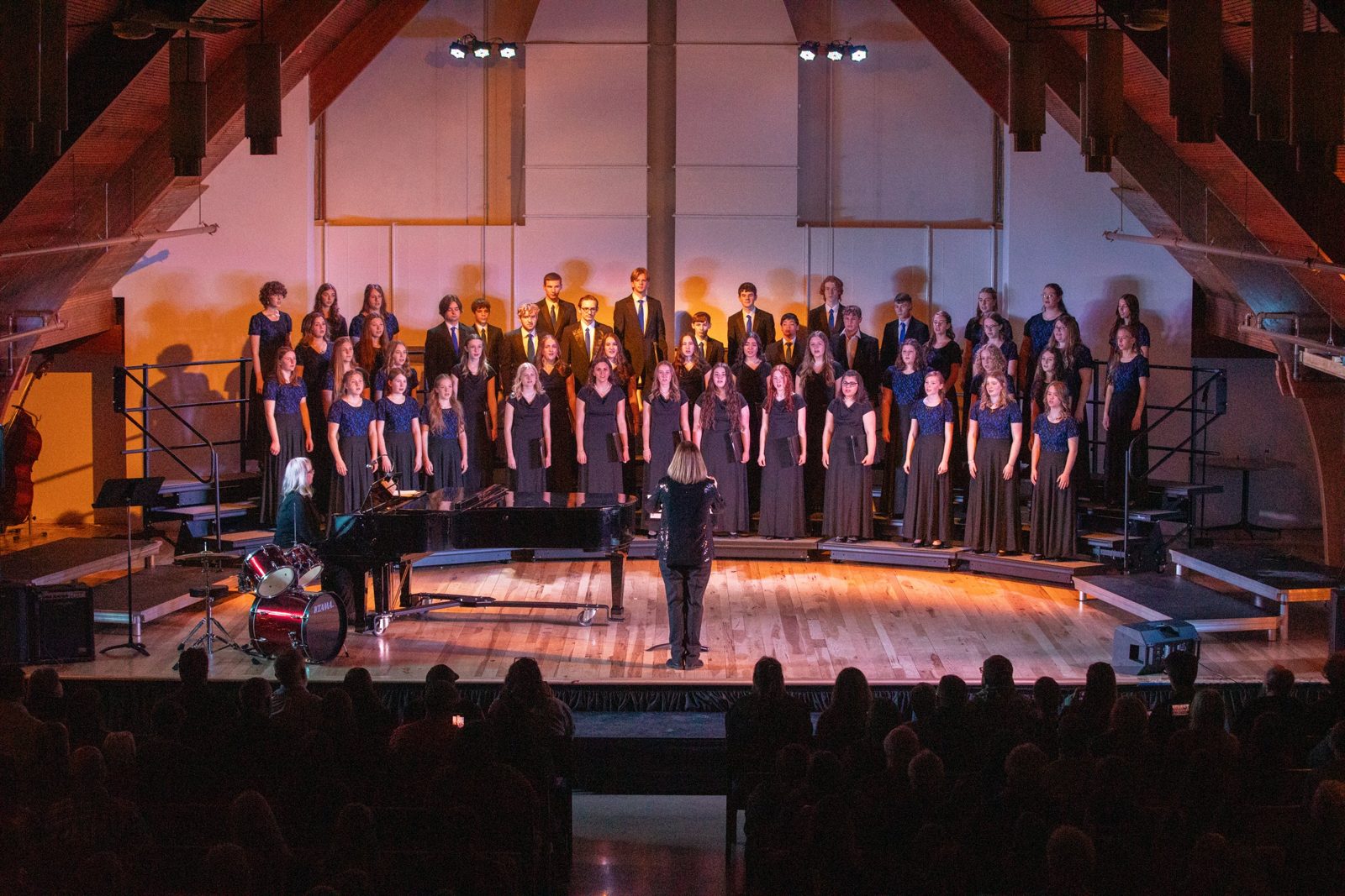 A choir in concert on a lit stage, Cathy conducting
